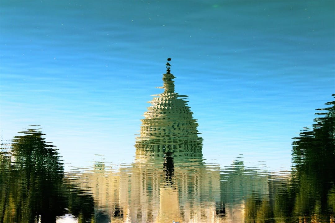 Reflection of a domed building and trees distorting on the surface of a blue, rippling body of water under clear sky.