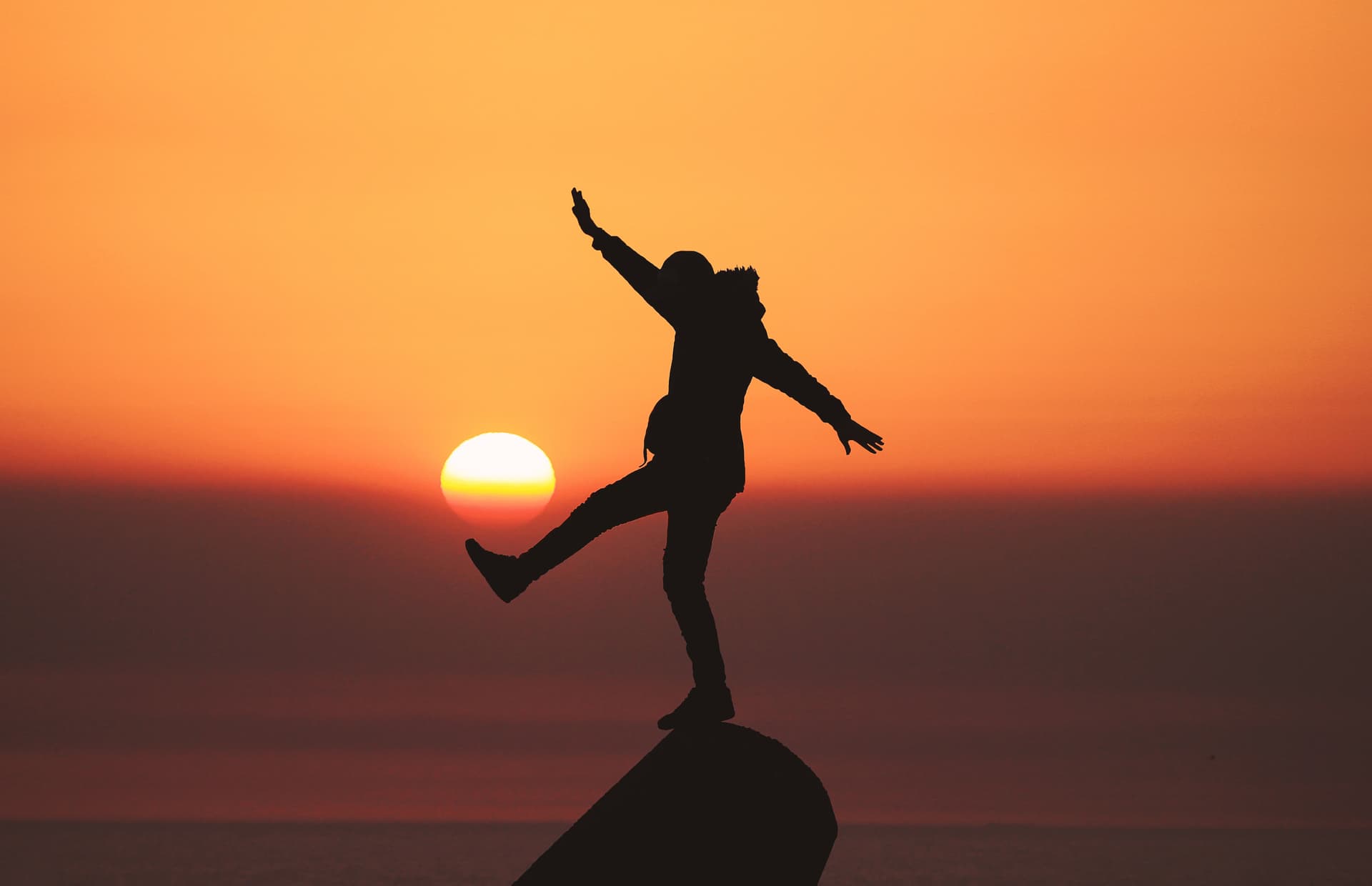 Silhouette of a person balancing on one leg atop a rock with arms outstretched, set against a vivid orange sunset with the sun low on the horizon.