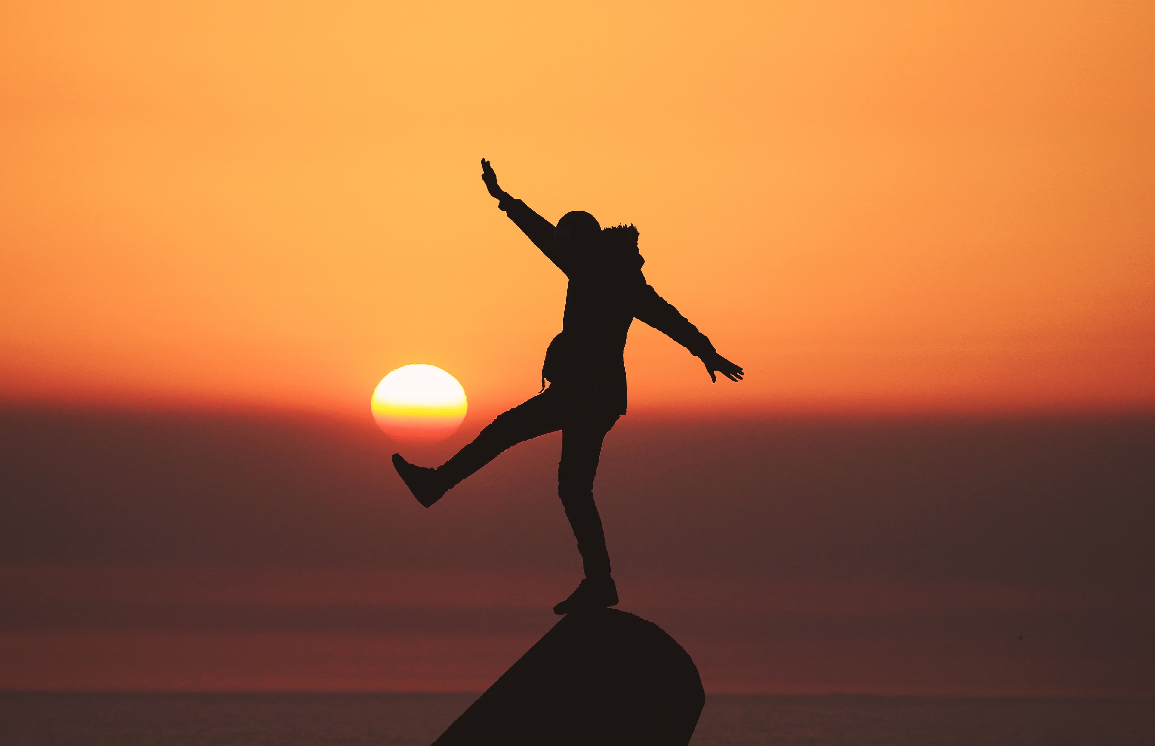 Silhouette of a person balancing on one leg atop a rock with arms outstretched, set against a vivid orange sunset with the sun low on the horizon.