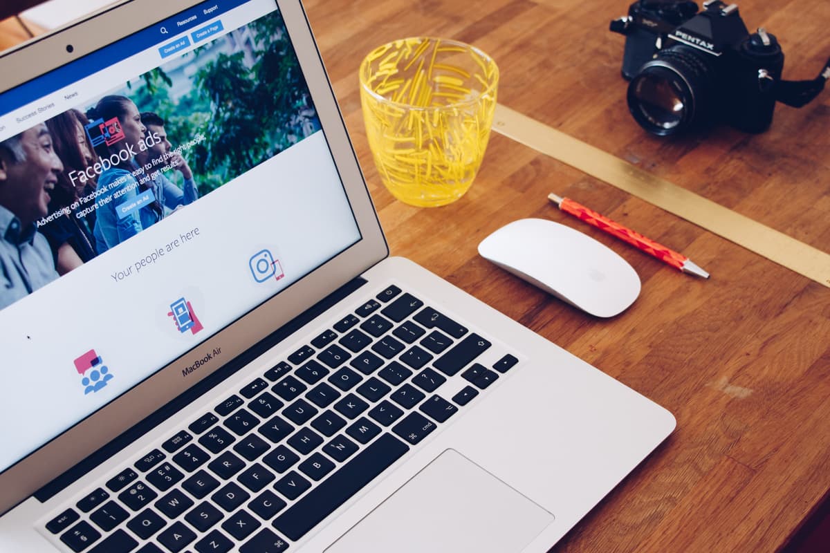 A MacBook Air displaying Facebook Ads on its screen sits on a wooden desk. Nearby are a wireless mouse, a yellow cup filled with paper clips, an orange pencil, and a Pentax camera, creating a workspace setup focused on digital marketing and advertising tools.